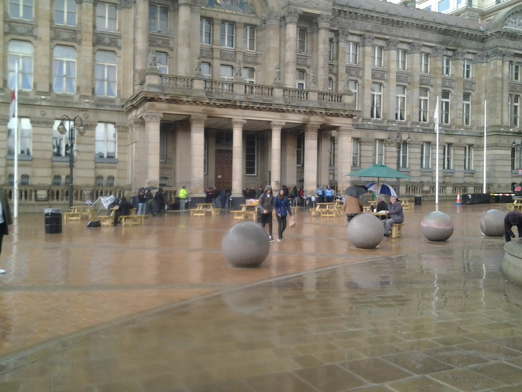 A wet Victoria Square with a few people walking towards the camera