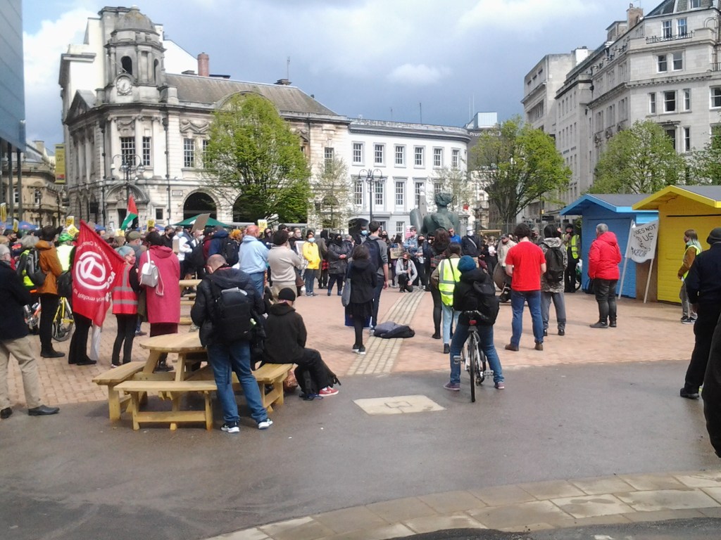 A crowd in Victoria Square, Birmingham. On the left woman with a large red flag. A Palestine flag is in the background.