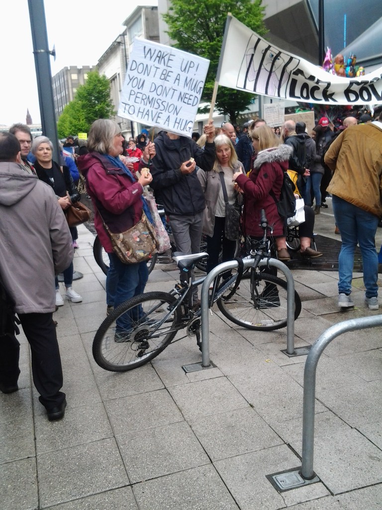 Woman holding sign "Wake Up! Don't be a mug. You don't need permission to have a hug." in a crowd of protesters