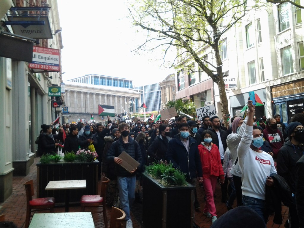 Large crowd of Palestine protesters marching through the city centre. Many in the front are wearing blue masks. Sign in the background says "Stop Funding Israel". 