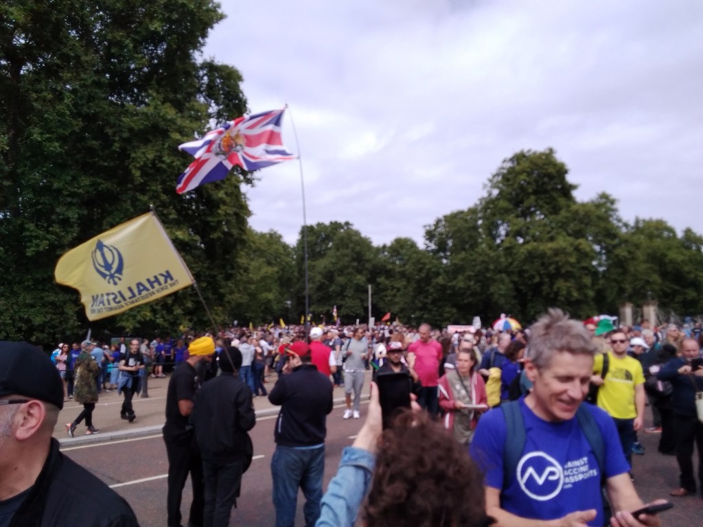 Man in front wearing a blue 'Against Vaccine Passports' T shirt, Sikh man wearing a turban waves a flag. Union Jack is present in the background.