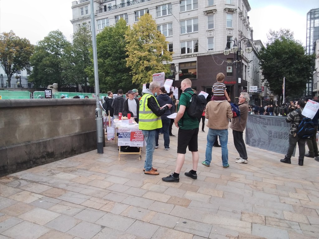 Stall by the Socialist party. Man in front wearing a hi viz jacket talking to another man wearing a green top. 