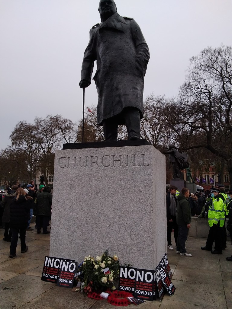 Statue of Winston Churchill on a plinth. Signs surrounding the plinth read 'NO to exclusion, to checkpoints, to discrimination, Covid ID'