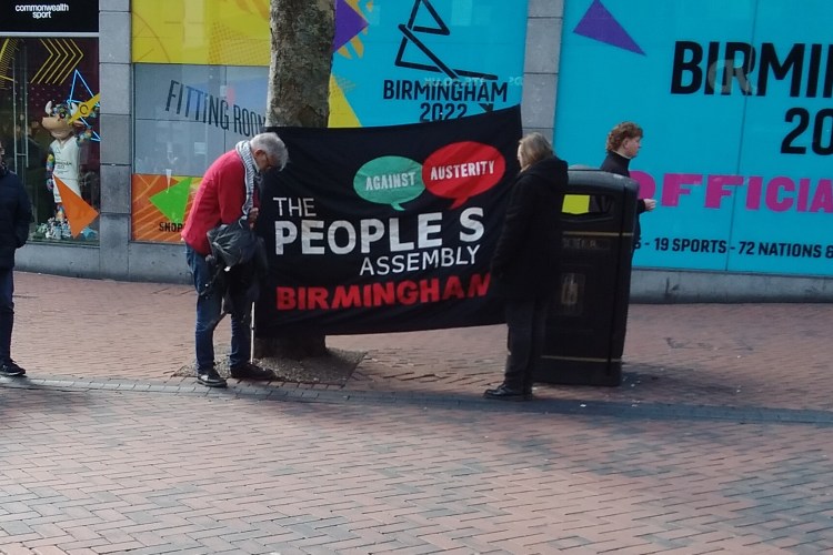 Black banner stating "The People's Assembly Birmingham: Against Austerity". Advertisements for Birmingham Commonwealth Games are in the background.