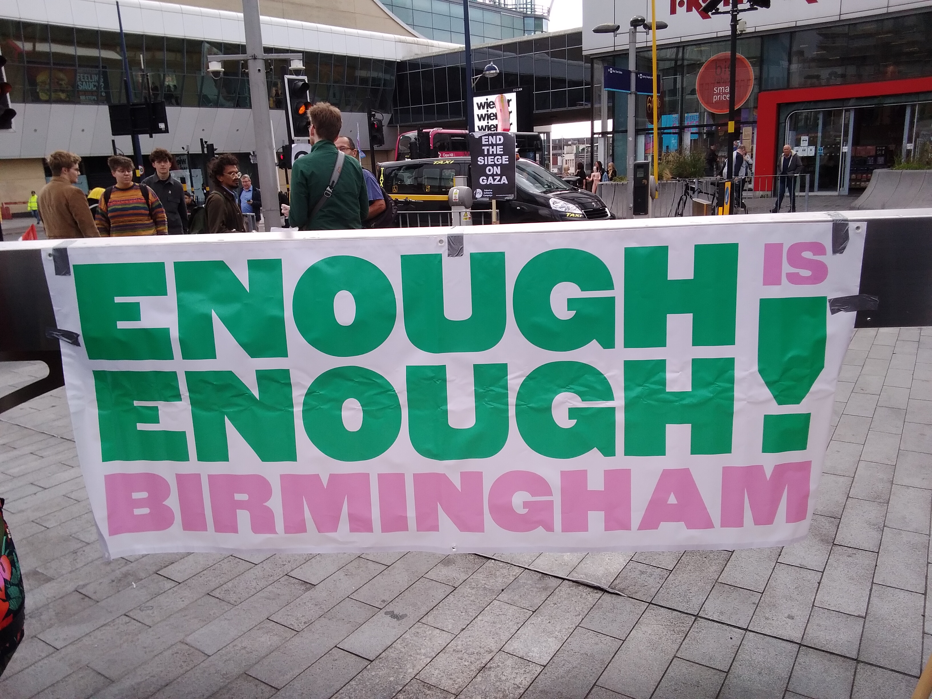 Banner reading Enough Is Enough Birmingham. Sign in the background reads 'End the Siege on Gaza'.