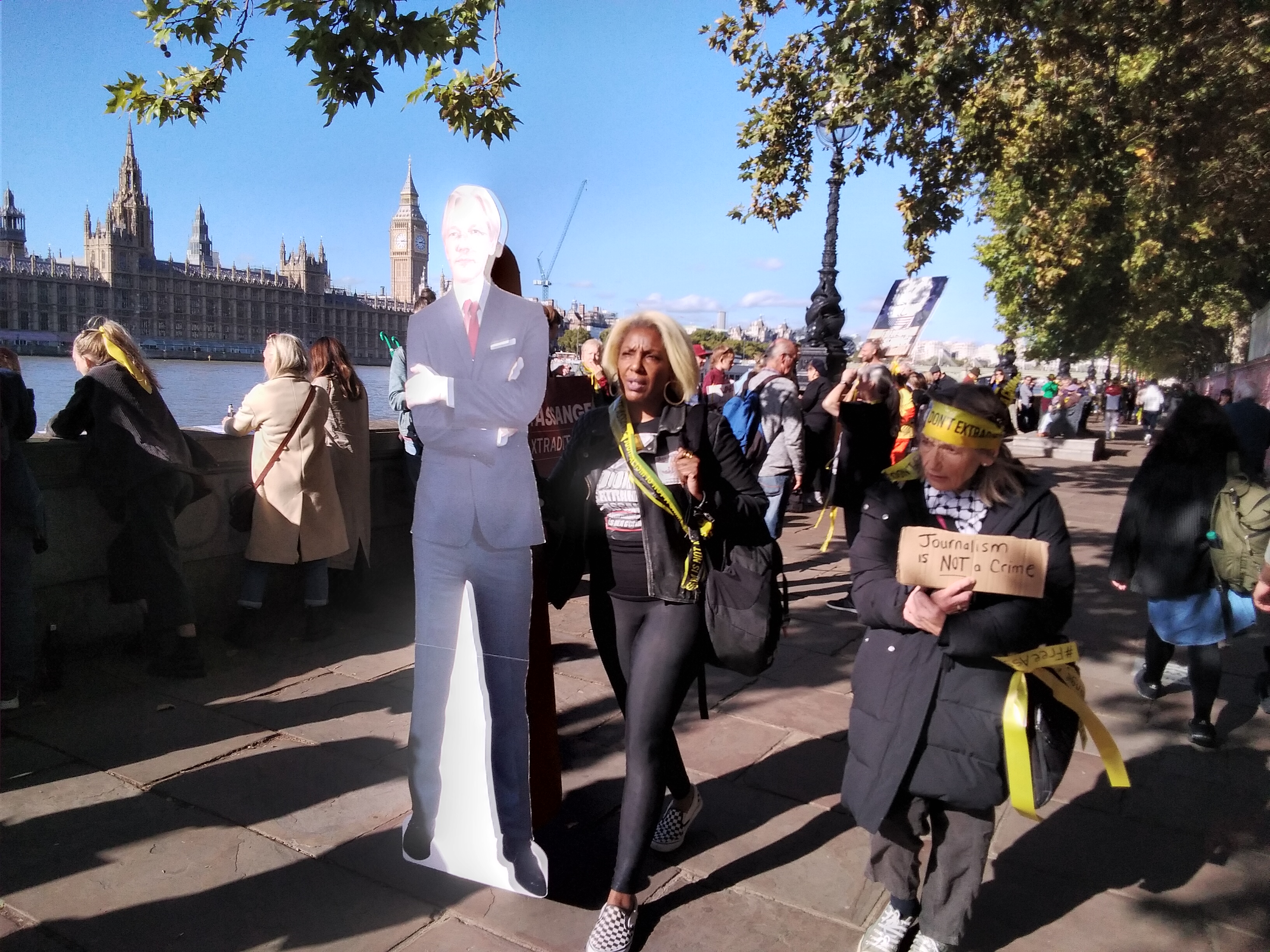 Woman carrying cardboard cutout of Julian Assange with crowds of people looking out over the Thames in the background.