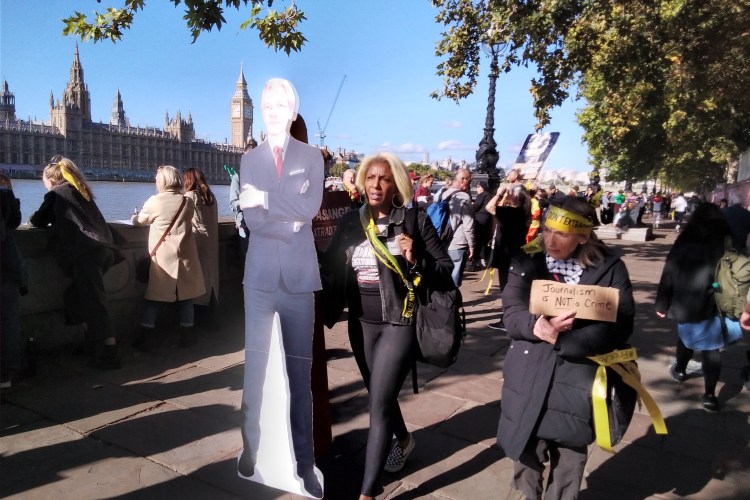Woman carrying cardboard cutout of Julian Assange with crowds of people looking out over the Thames in the background.