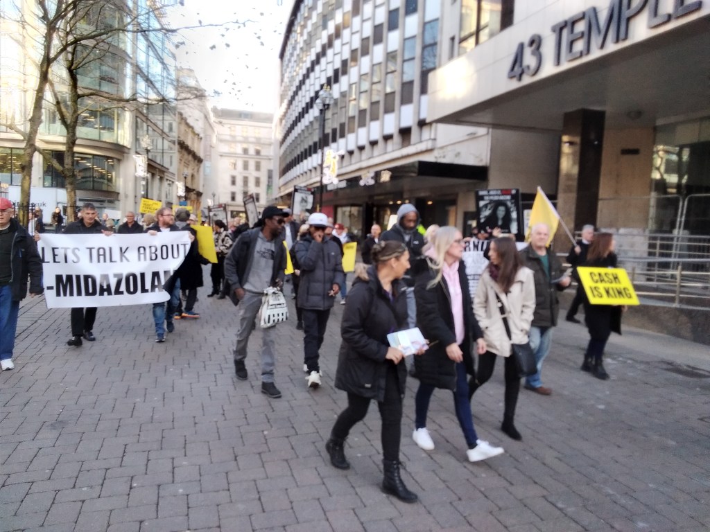 Sign reading "Let's talk about Midazolam" among a marching crowd.
