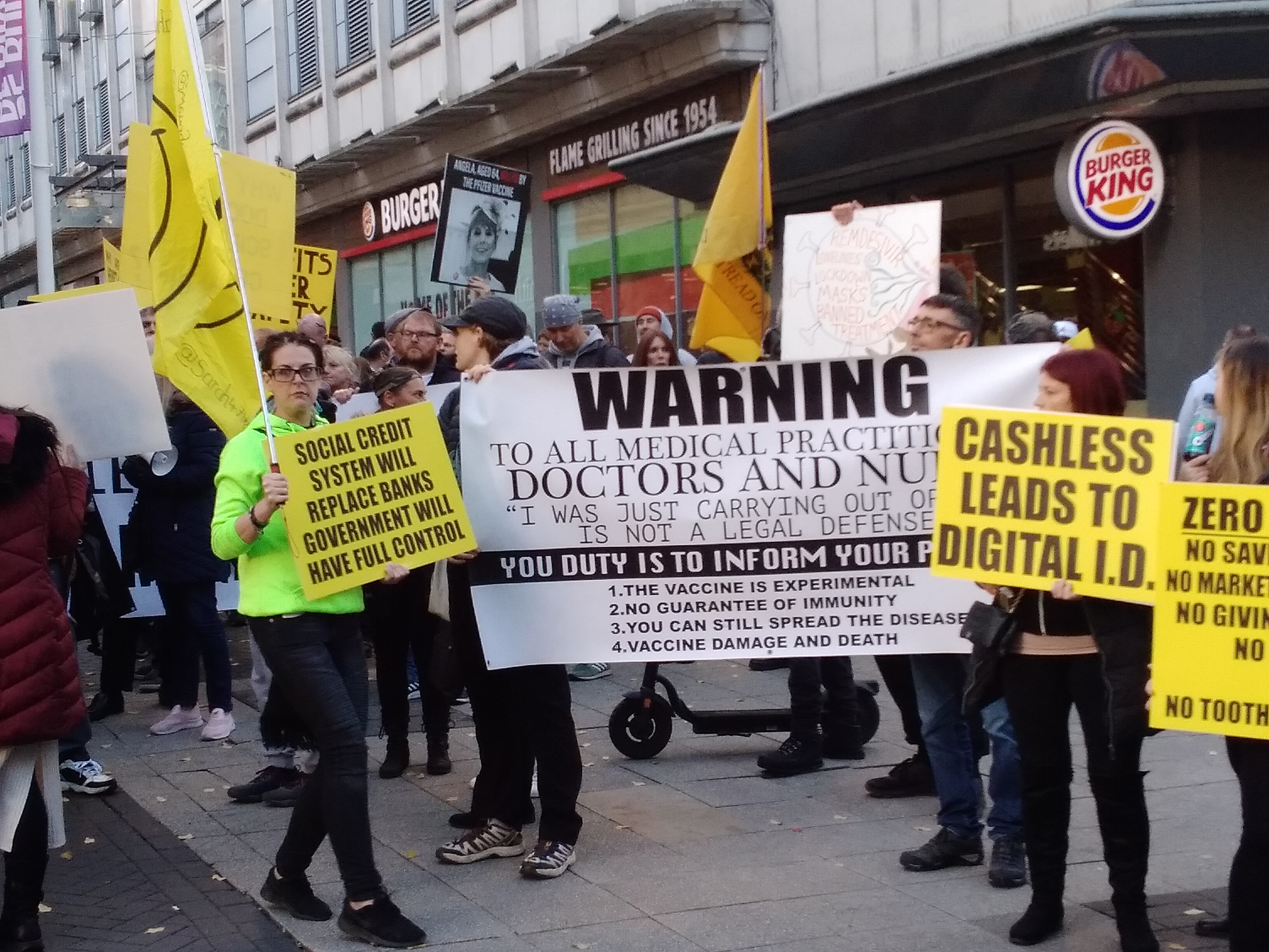 Crowd with yellow flags and signs reading 'Cashless leads to Digital ID' 'Social credit system will replace banks' and a sign reminded doctors and nurses they need to give informed consent.