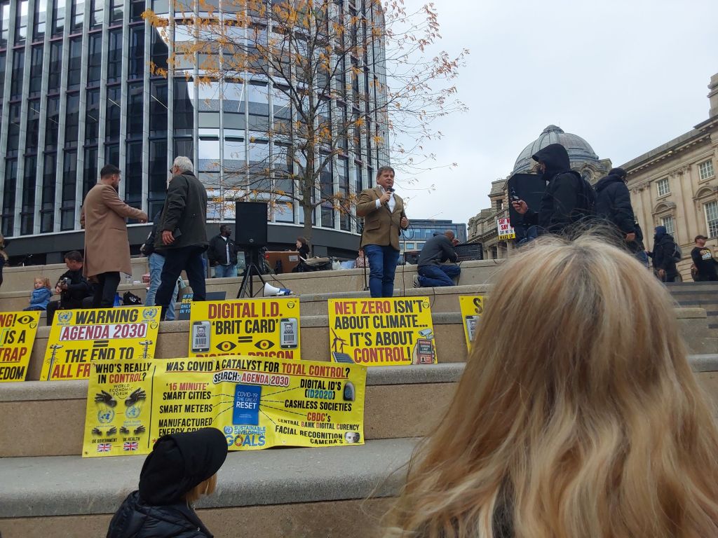 MP Andrew Bridgen stands at the top of the stairs. Multiple yellow signs with text criticising digital ID, agenda 2030 and net zero and below him.