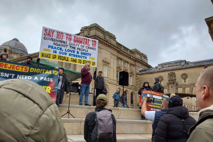 Man in a maroon shirt stands in front of a banner reading 'Say No to Digital ID'